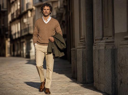 Mann in weißem Hemd mit karamellfarbenem Merino-Pullover und olivgrüner Übergangsjacke über dem Arm geht durch eine morgendliche Straße — Layering-Outfit für die Übergangszeit zwischen Sommer und Herbst aus der Huberd Kollektion